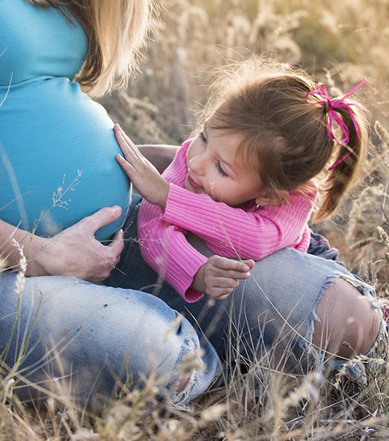Mother and child touching pregnant belly while sitting in a field.